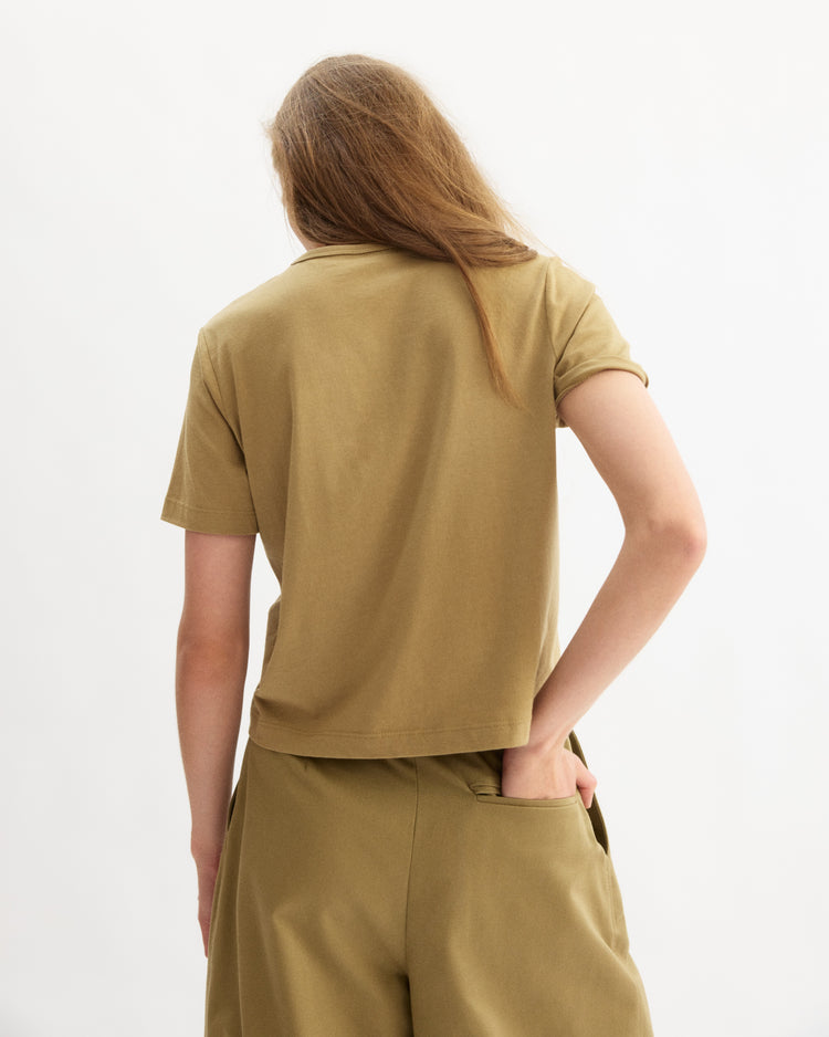 A person with long, light brown hair stands with their back to the camera wearing the Arlo Mott Willow Short Sleeve Tee in Khaki and matching pants, styled effortlessly against a plain background.