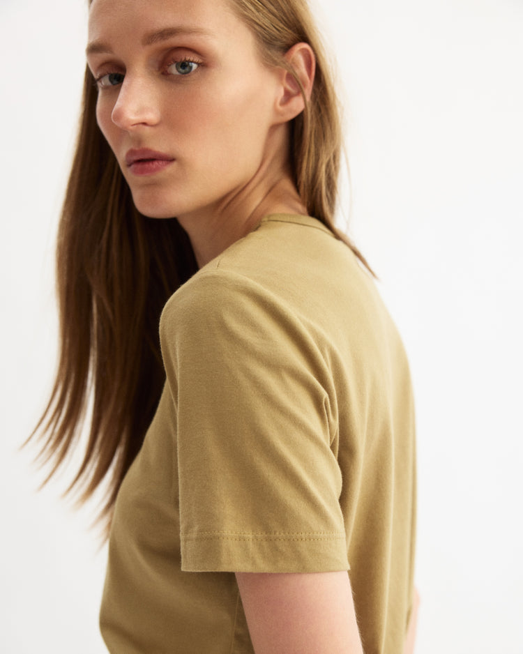 A person with long, straight light brown hair wears the Arlo Mott Willow Short Sleeve Tee in Khaki, looking over their shoulder at the camera against a plain white background.