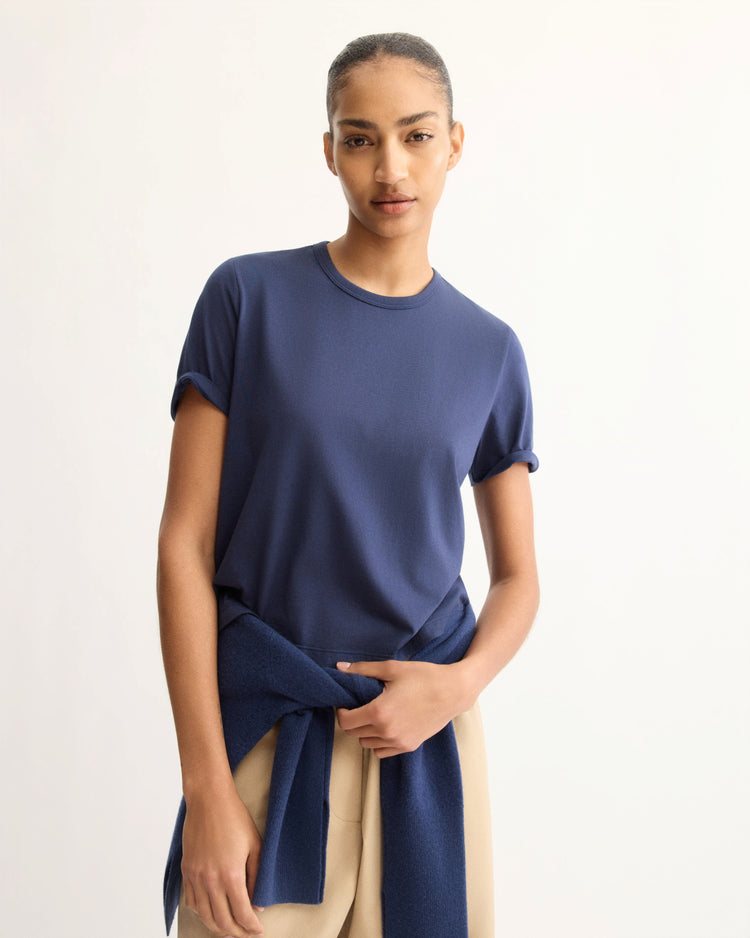 A person wears the Arlo Mott Willow Short Sleeve Tee in Navy with beige pants, standing against a plain light background, and has a matching navy sweater tied around their waist.
