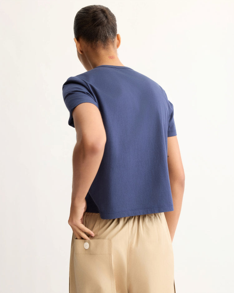 A person, hair tied back, faces away against a plain light background wearing the Arlo Mott Willow Short Sleeve Tee in Navy and beige pants with a buttoned pocket, their right hand resting on their hip.