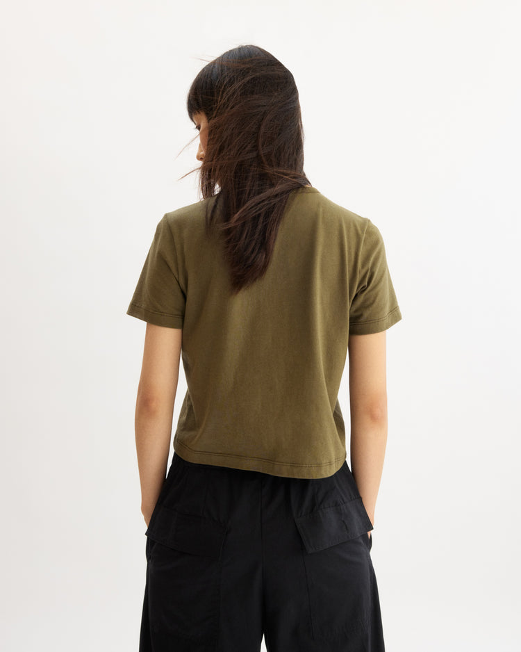 A person with long dark hair, facing away, wears the Arlo Mott Willow Short Sleeve Tee in Olive (boxy fit, made in New York) with black pants against a plain white background.