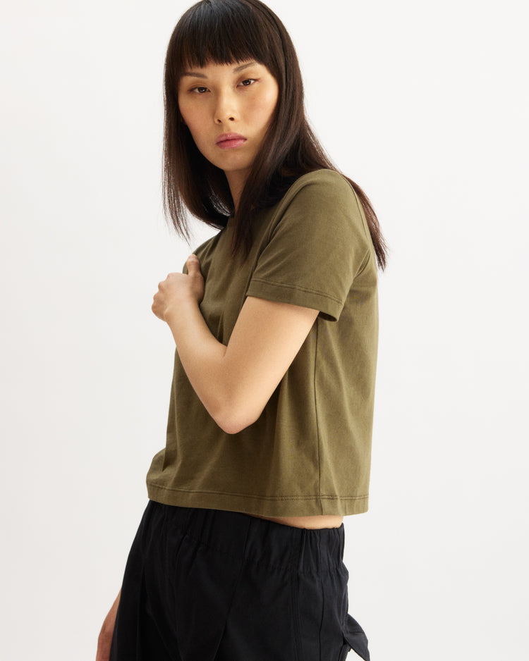 A person with straight, dark brown hair and bangs is wearing the Arlo Mott Willow Short Sleeve Tee in Olive and black pants, standing against a white background and looking slightly to the side.