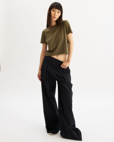 Against a plain white backdrop, a person wears the Arlo Mott Willow Short Sleeve Tee in Olive with wide-leg black pants and black shoes, posing with one hand on their hip and the other at their side.