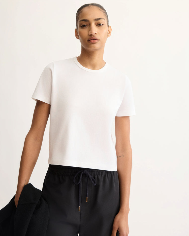 A person with slicked-back hair stands against a light background wearing the Arlo Mott Willow Short Sleeve Tee in White and black drawstring pants, holding a black garment in one hand.