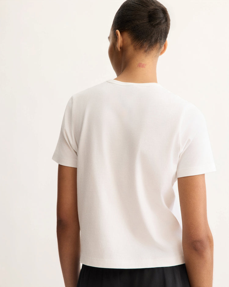 Seen from behind, a person with short hair wears the Arlo Mott Willow Short Sleeve Tee in White and black pants. A small red tattoo appears on their neck against a plain, light-colored background.