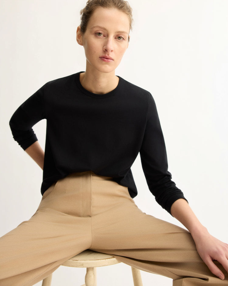 A person with light skin and tied-back hair sits on a stool against a plain backdrop, wearing the Arlo Mott Sullivan Long Sleeve Tee in Black and high-waisted beige trousers, looking directly at the camera with a neutral expression.