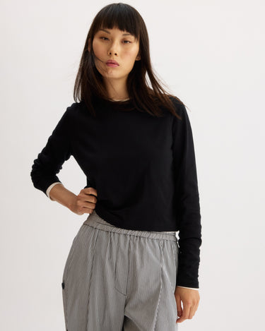 A woman with straight dark hair and bangs wears the Arlo Mott Sullivan Long Sleeve Tee in Black and high-waisted, loose-fitting, light gray pinstriped pants, standing against a plain light background with a neutral expression.
