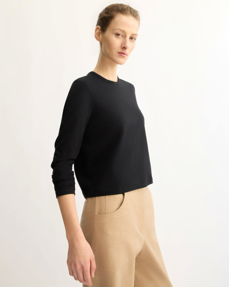 A person with light skin and tied-back hair wears the Arlo Mott Sullivan Long Sleeve Tee in Black with high-waisted beige pants, standing against a plain light background. They have a neutral expression and are facing slightly to the side.