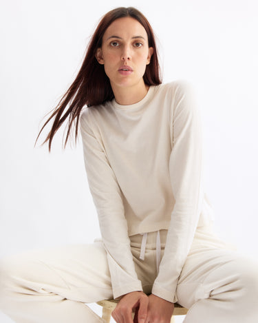 A woman with long brown hair sits on a stool, wearing the Arlo Mott Sullivan Long Sleeve Tee in Chalk with matching pants, looking directly at the camera against a plain white background.