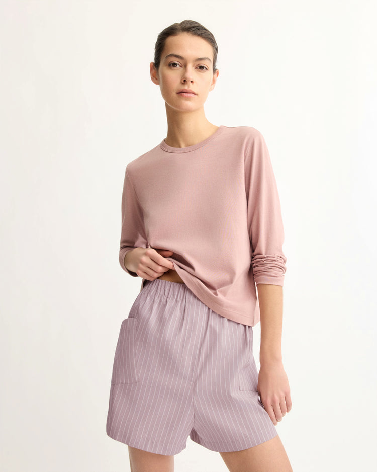 A person wearing the Arlo Mott Sullivan Long Sleeve Tee in Dusty Rose and loose, high-waisted pink pinstriped shorts stands against a plain, light background, facing the camera.