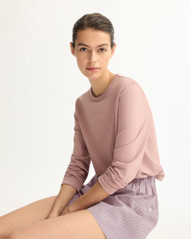 A person with dark hair sits looking at the camera, wearing the Arlo Mott Sullivan Long Sleeve Tee in Dusty Rose and light purple pinstriped shorts, against a plain light background.