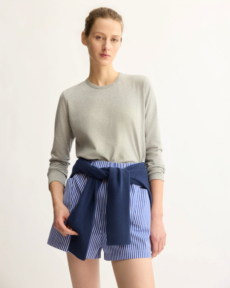 A person wearing the Arlo Mott Sullivan Long Sleeve Tee in Heather Grey, blue and white striped shorts, and a navy sweater tied around their waist stands against a plain light background.