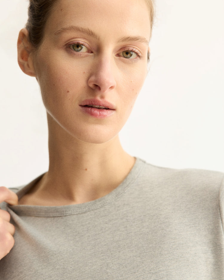 A woman with light skin and pulled-back hair wears the Arlo Mott Sullivan Long Sleeve Tee in Heather Grey, gently holding the neckline with one hand as she gazes softly at the camera against a plain background.