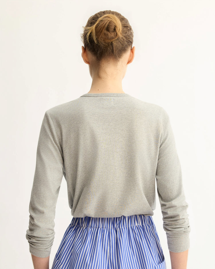 A person with light brown hair in a bun, shown from behind against a plain light background, is wearing the Arlo Mott Sullivan Long Sleeve Tee in Heather Grey and a blue-and-white vertically striped skirt.
