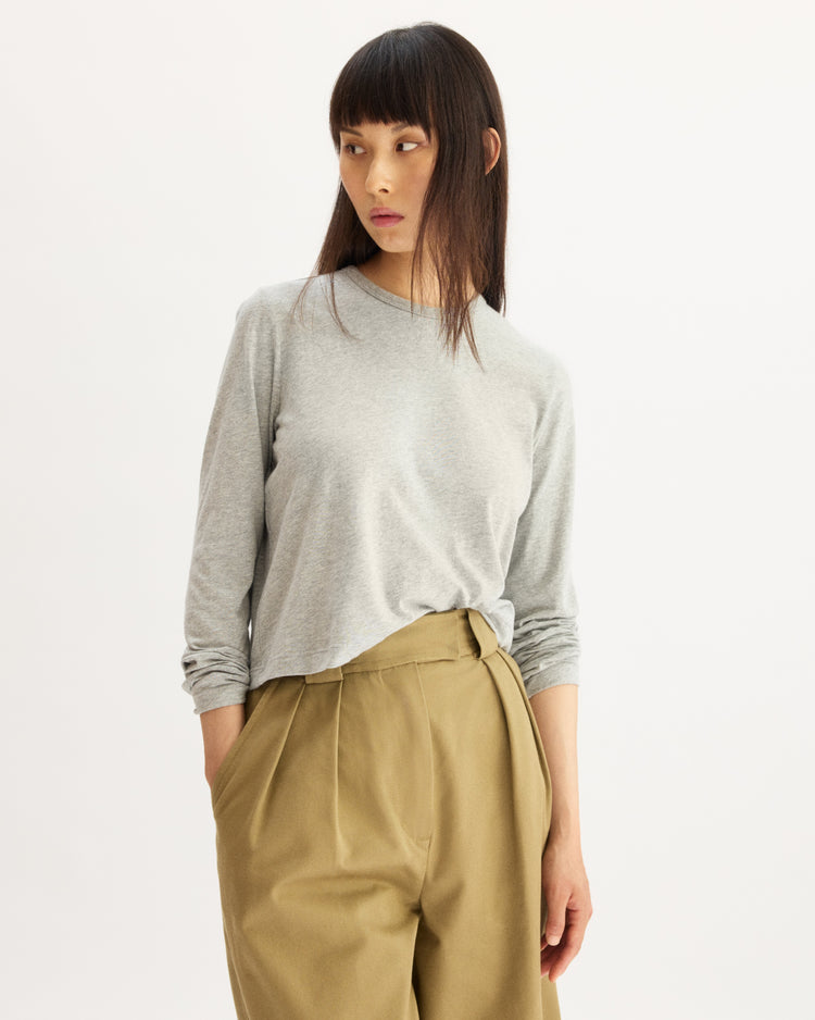 A woman in an Arlo Mott Sullivan Long Sleeve Tee in Heather Grey, tucked into high-waisted khaki pants, stands against a plain white background and looks to her left with a neutral expression.