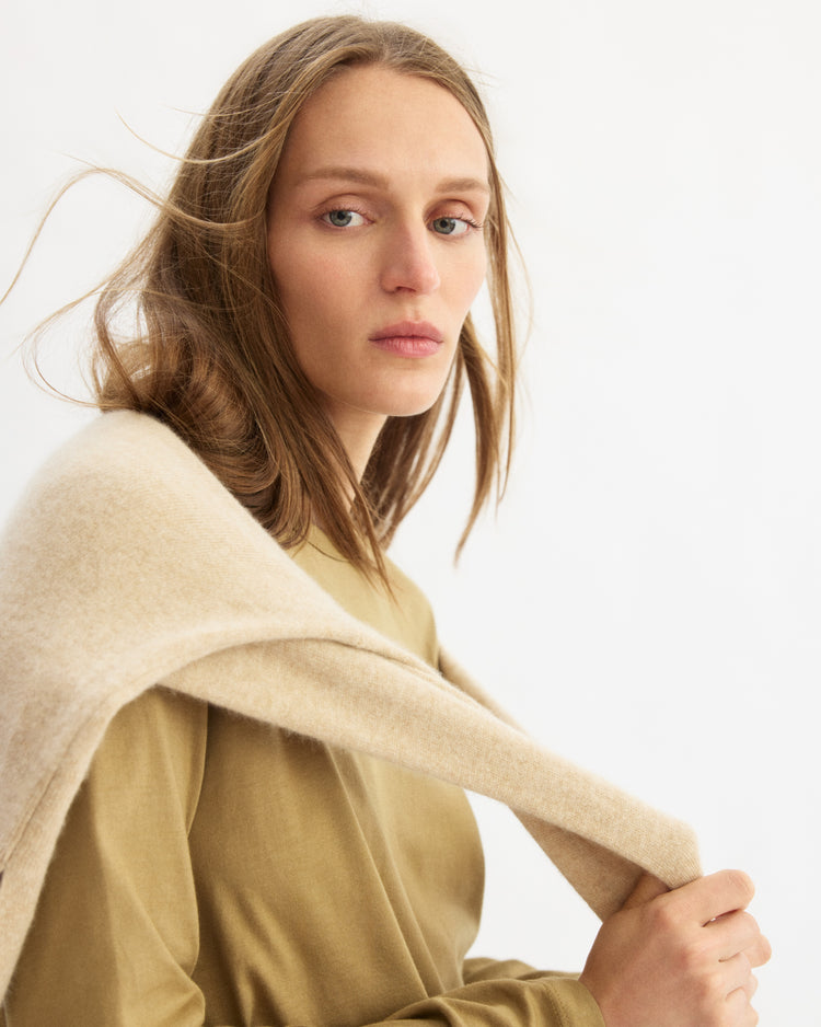 A woman with light brown hair and a neutral expression faces the camera, wearing a beige wool sweater over the Arlo Mott Sullivan Long Sleeve Tee in Khaki. The background is plain white.