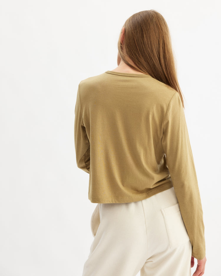 A person with long light brown hair stands with their back to the camera against a white background, wearing Arlo Mott’s Sullivan Long Sleeve Tee in Khaki and cream-colored pants.