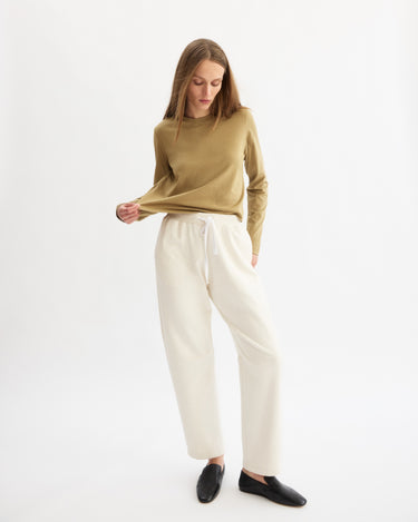 A woman stands against a white background, wearing the Arlo Mott Sullivan Long Sleeve Tee in Khaki with white drawstring pants and black slip-on shoes. She looks down, gently pulling at her shirt with one hand.