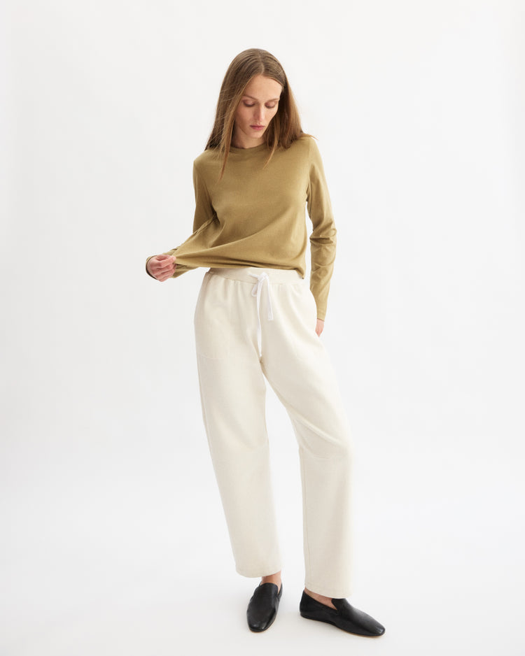 A woman stands against a white background, wearing the Arlo Mott Sullivan Long Sleeve Tee in Khaki with white drawstring pants and black slip-on shoes. She looks down, gently pulling at her shirt with one hand.