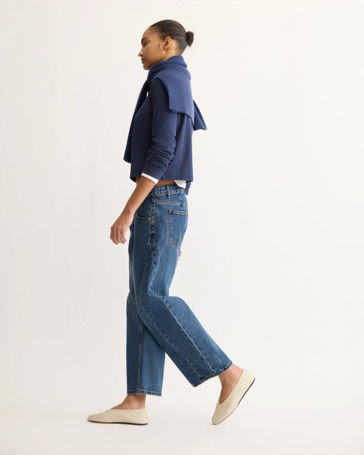 A person with hair in a bun wears an Arlo Mott Sullivan Long Sleeve Tee in Navy draped over their shoulders, a boxy Supima cotton T-shirt, blue jeans, and cream flats, walking against a plain light background.