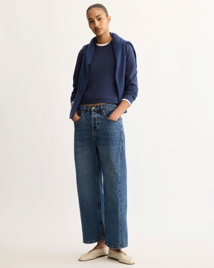 A woman stands with hands in her pockets, wearing the Arlo Mott Sullivan Long Sleeve Tee in Navy, a matching sweater over her shoulders, wide-leg blue jeans, and cream-colored shoes against a plain light background.