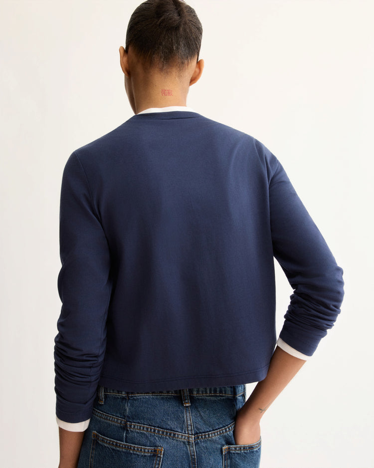 A person with short hair stands with their back to the camera, wearing Arlo Mott's Sullivan Long Sleeve Tee in Navy and blue jeans. Their left hand is on their hip, set against a plain off-white wall.