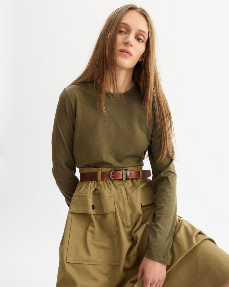 A young woman with long, straight light brown hair wears the Arlo Mott Sullivan Long Sleeve Tee in Olive, tucked into high-waisted khaki pants with a brown belt, standing against a plain white background.