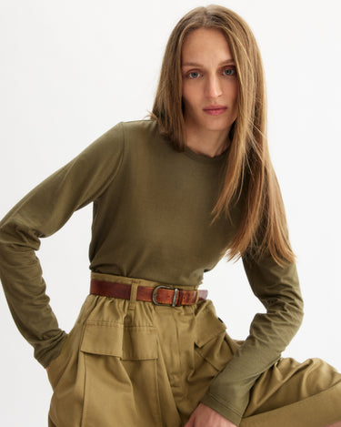 A woman with long straight hair models the Arlo Mott Sullivan Long Sleeve Tee in Olive, tucked into high-waisted khaki pants with large pockets and a brown belt, posing against a plain white background.