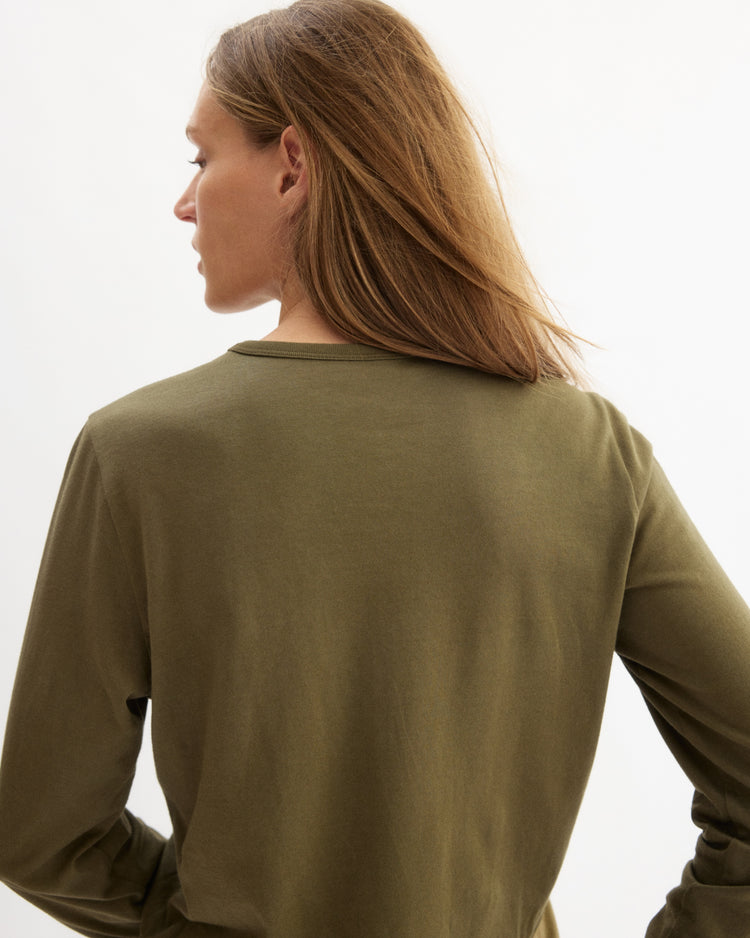 A person with long, light brown hair is seen from behind, wearing an olive Sullivan Long Sleeve Tee by Arlo Mott against a plain light background.