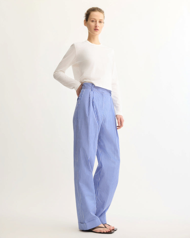 A person stands against a plain background wearing a white long-sleeve shirt, Arlo Mott Spencer Pleated Pants in Blue Stripe, and black sandals.