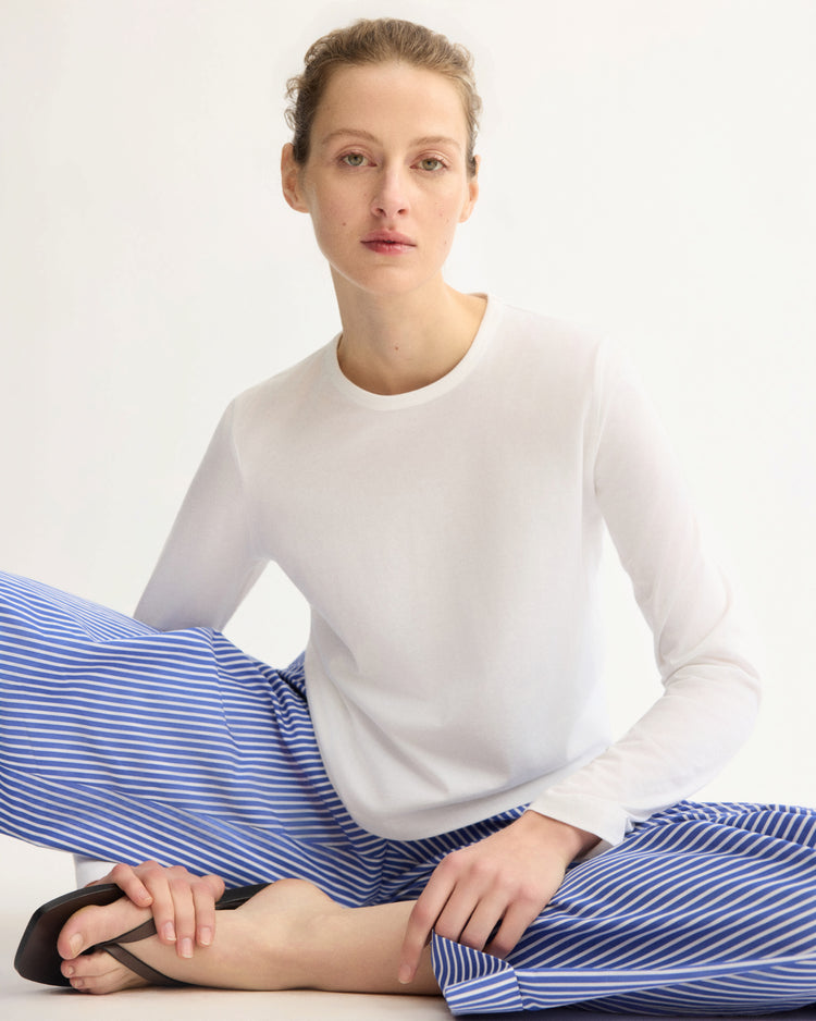 A person with light skin and tied-back hair sits on the floor wearing the Arlo Mott Sullivan Long Sleeve Tee in White and blue-and-white striped pants, looking directly at the camera against a plain background.