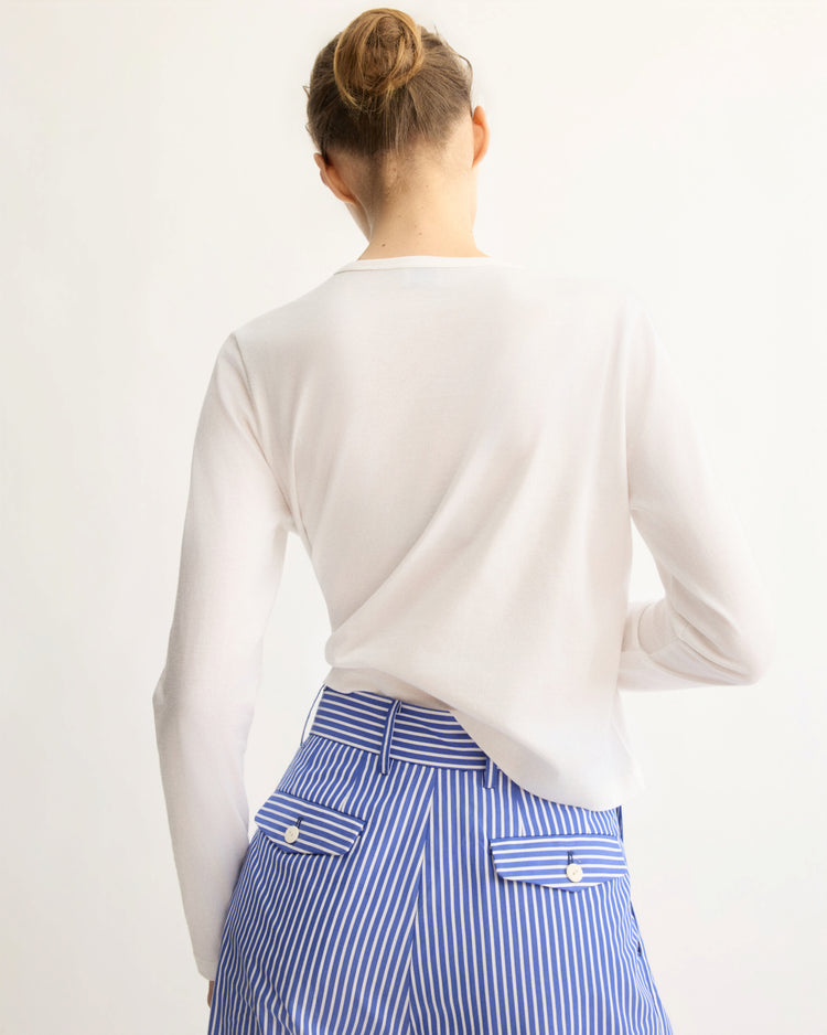A person with hair in a neat bun is seen from behind, wearing the Arlo Mott Sullivan Long Sleeve Tee in White and high-waisted blue and white vertically striped pants with back pockets.