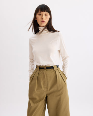 A woman with straight dark hair and a neutral expression stands against a plain background, wearing the Arlo Mott Prince Long Sleeve Turtleneck in Chalk tucked into high-waisted, wide-leg khaki pants with her hands in her pockets.