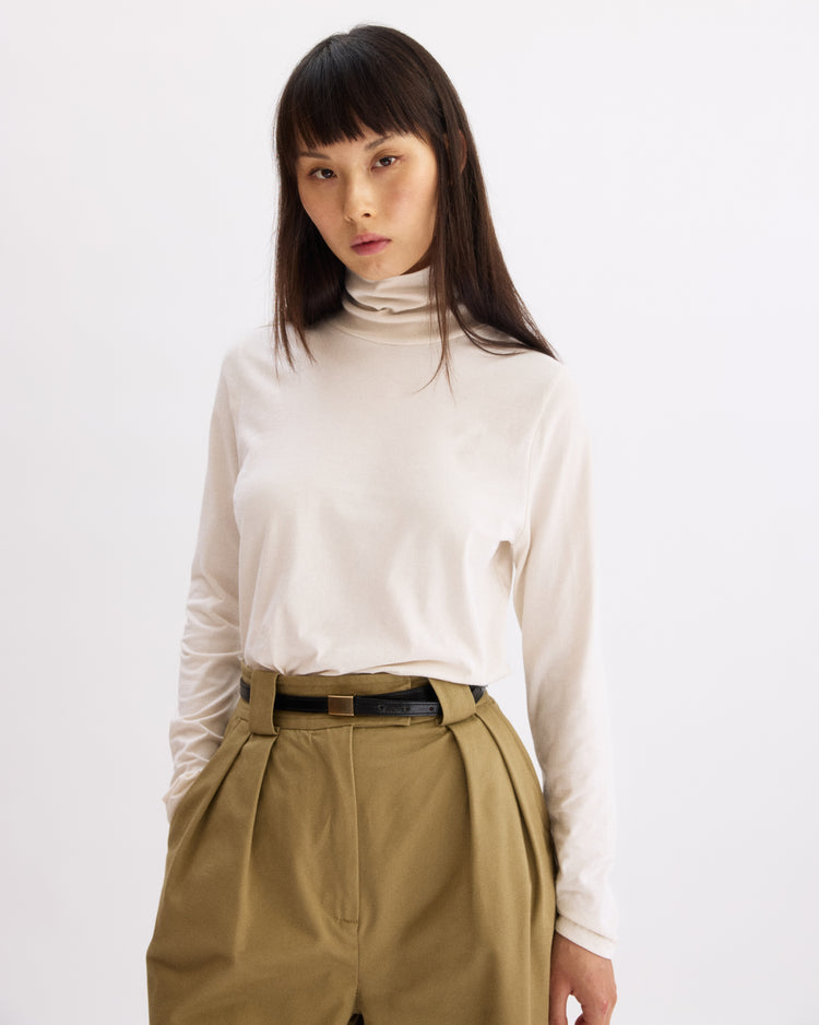 A person with straight dark hair and bangs wears the Arlo Mott Prince Long Sleeve Turtleneck in Chalk, tucked into high-waisted pleated khaki pants with a black belt, standing against a plain white background.