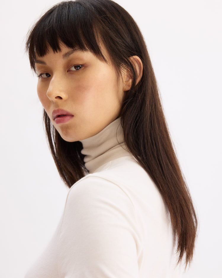 A woman with long straight dark hair and blunt bangs wears the Arlo Mott Prince Long Sleeve Turtleneck in Chalk, gazing sideways with a neutral expression against a plain light background.