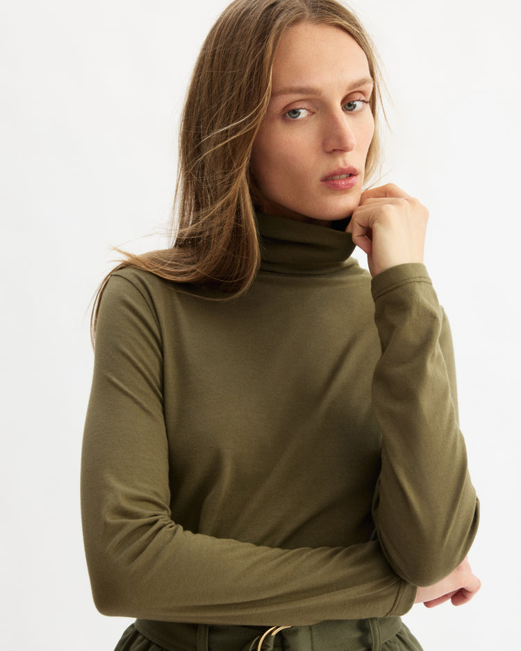 A woman with long, light brown hair models the Arlo Mott Prince Long Sleeve Turtleneck in Olive against a white background, her left hand touching her chin as she looks at the camera—simple and chic Brooklyn style.