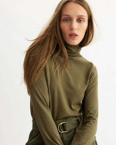 A woman with long brown hair wears the Arlo Mott Prince Long Sleeve Turtleneck in Olive, paired with matching belted pants. She stands against a white background with a neutral expression, her hair tousled for effortless Brooklyn style.