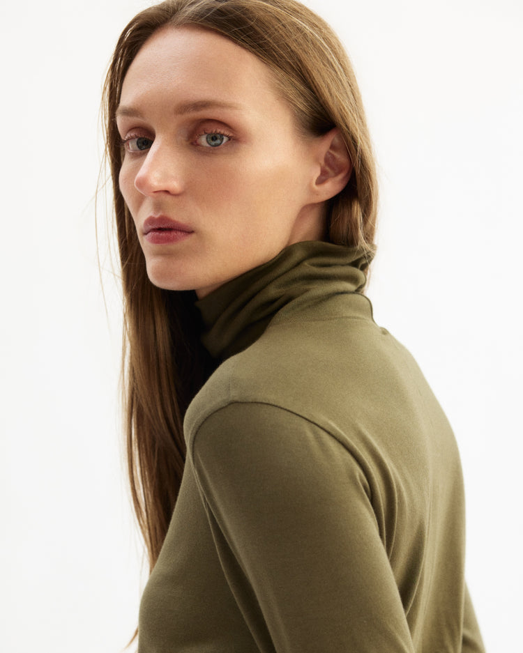 A person with long straight brown hair models the Arlo Mott Prince Long Sleeve Turtleneck in Olive, glancing over their shoulder against a light background and embodying relaxed Brooklyn style.