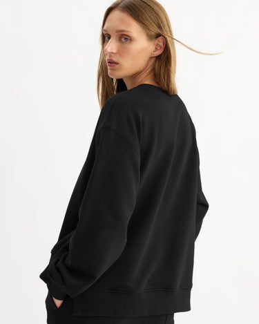 A person with long, light brown hair stands sideways against a plain white background, looking back over their shoulder while wearing the Arlo Mott Delancey Crew Neck Sweatshirt in Black.