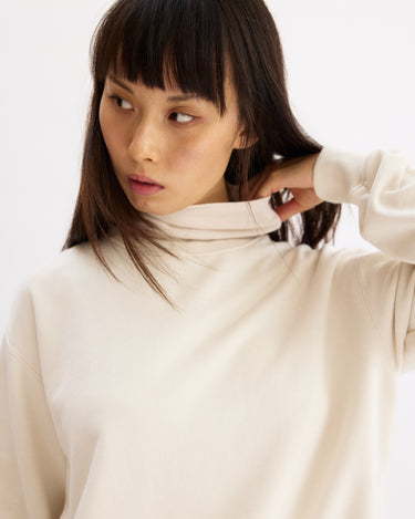A woman with straight dark hair and bangs wears the Arlo Mott Delancey Crew Neck Sweatshirt in Chalk, looking to her left as she gently adjusts the collar. The background is plain and light.