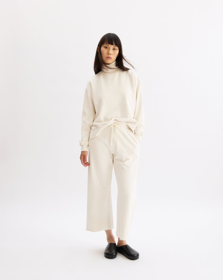 A woman with straight dark hair and bangs wears the Arlo Mott Delancey Crew Neck Sweatshirt in Chalk, styled as a modern sweatsuit with matching wide-leg pants and black slip-on shoes, standing against a plain white background.