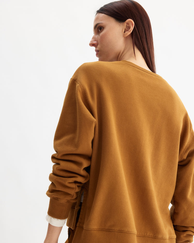 A woman with straight brown hair is shown from the back and side, looking over her shoulder while wearing the Arlo Mott Delancey Crew Neck Sweatshirt in Cinnamon against a plain white background.