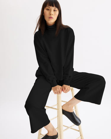 A woman with straight dark hair and bangs sits on a wooden stool against a plain white background, wearing Arlo Mott’s Kane Cropped Wide Sweatpants in Black, a black turtleneck, and black slip-on shoes.