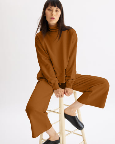 A woman with straight dark hair sits on a stool against a white background, wearing the Arlo Mott Delancey Crew Neck Sweatshirt in Cinnamon along with matching sweatsuit pants and black slip-on shoes.