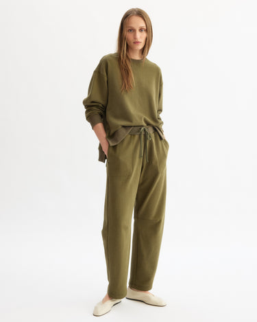 A woman with long, straight hair stands against a white background wearing the Arlo Mott Delancey Crew Neck Sweatshirt in Olive and matching drawstring pants, both in soft Italian terry cloth. She pairs the look with cream-colored slip-on shoes.