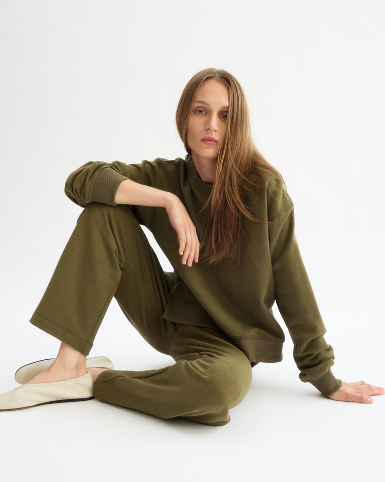 A woman with long brown hair sits on the floor against a white background, wearing the Arlo Mott Delancey Crew Neck Sweatshirt in Olive with matching pants and white slip-on shoes.