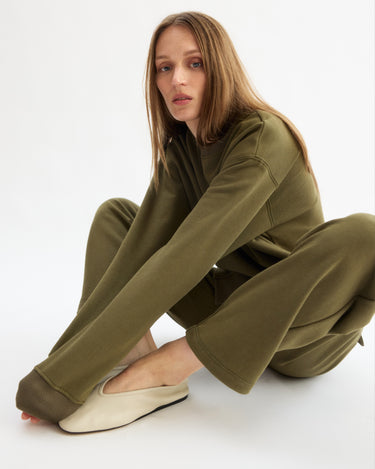 A person with long light brown hair wears the Arlo Mott Delancey Crew Neck Sweatshirt in Olive, sitting on the floor with one knee up and looking at the camera. They are barefoot in cream slip-on shoes against a white background.
