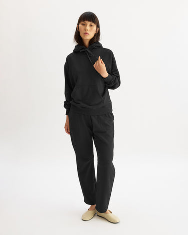 A woman with straight dark hair stands against a white background, wearing Arlo Mott's Baltic Barrel Leg Sweatpants in Black with a black hoodie and light beige slippers. One hand is raised near her collar.