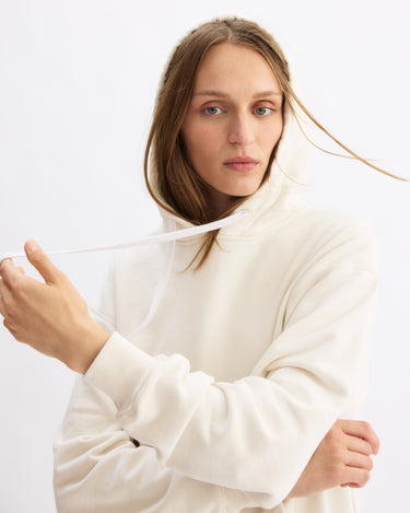 A woman with long, light brown hair wears the Arlo Mott Windsor Hooded Sweatshirt in Chalk, made from cozy Italian terry cloth. She holds one drawstring with her left hand and looks at the camera against a plain white background.