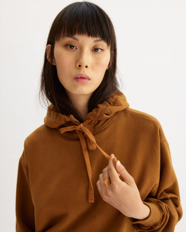 A person with straight dark hair and bangs wears the Arlo Mott Windsor Hooded Sweatshirt in Cinnamon, holding a drawstring in their right hand, standing against a plain light background.